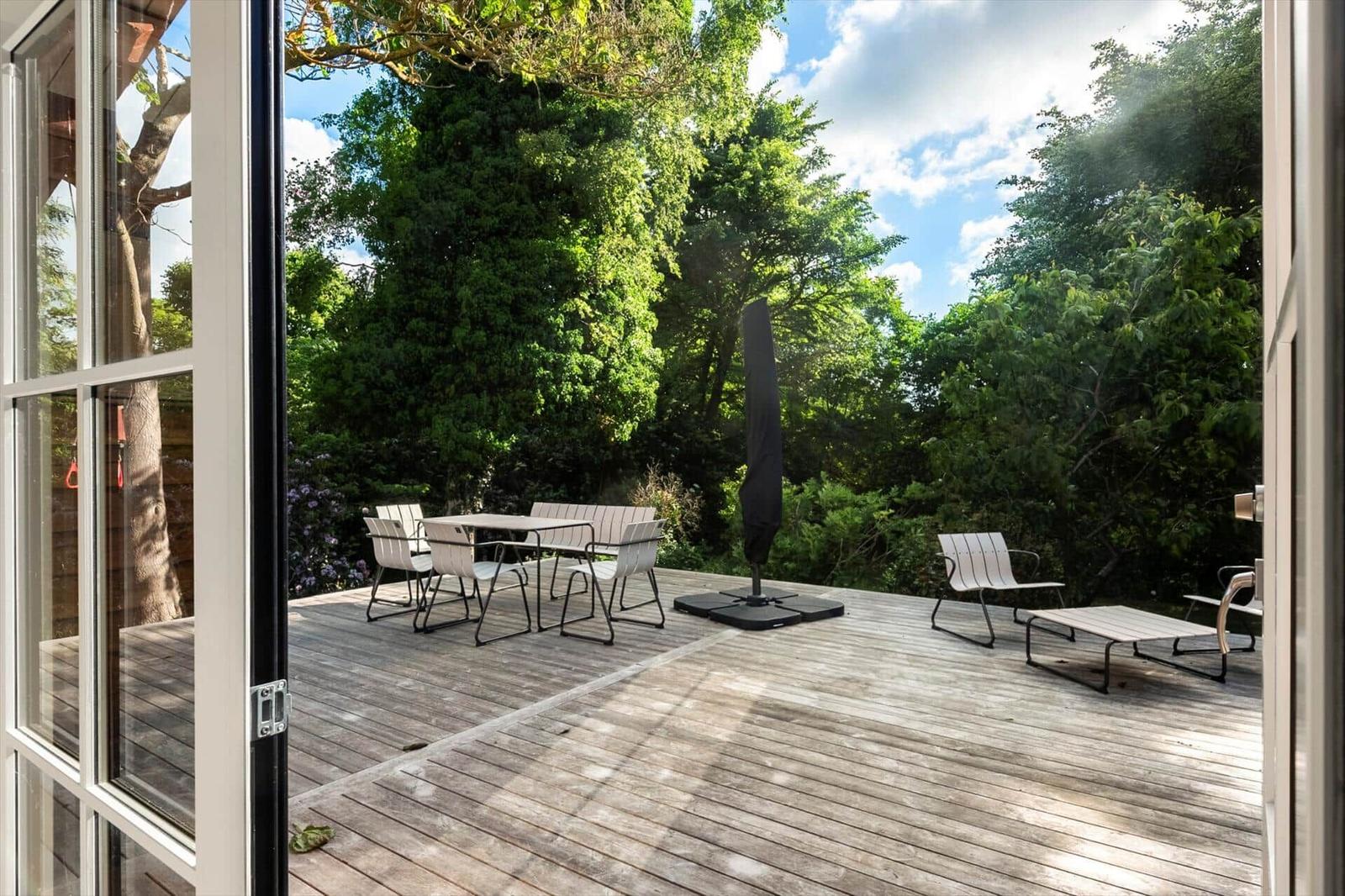 Terrasse mit Tisch, Stühlen und Sonnenschirm. Umgeben von Bäumen und Grünflächen.