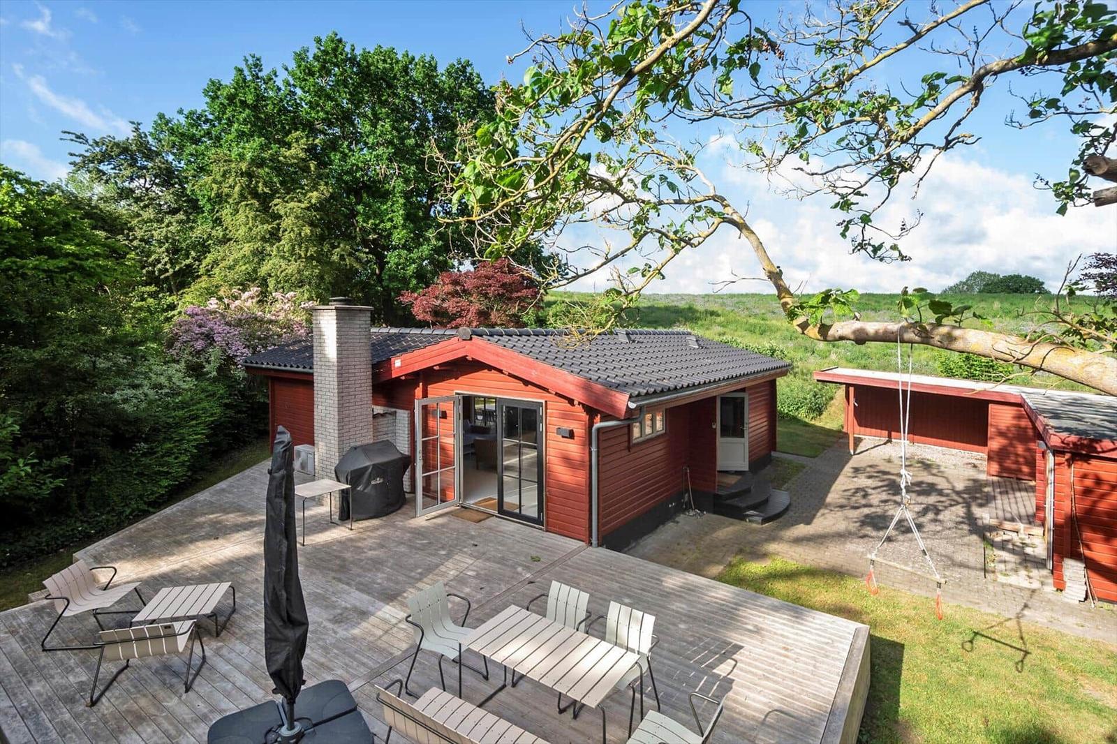 Rotes Holzhaus mit Terrasse, Grill und Schaukel im Grünen.