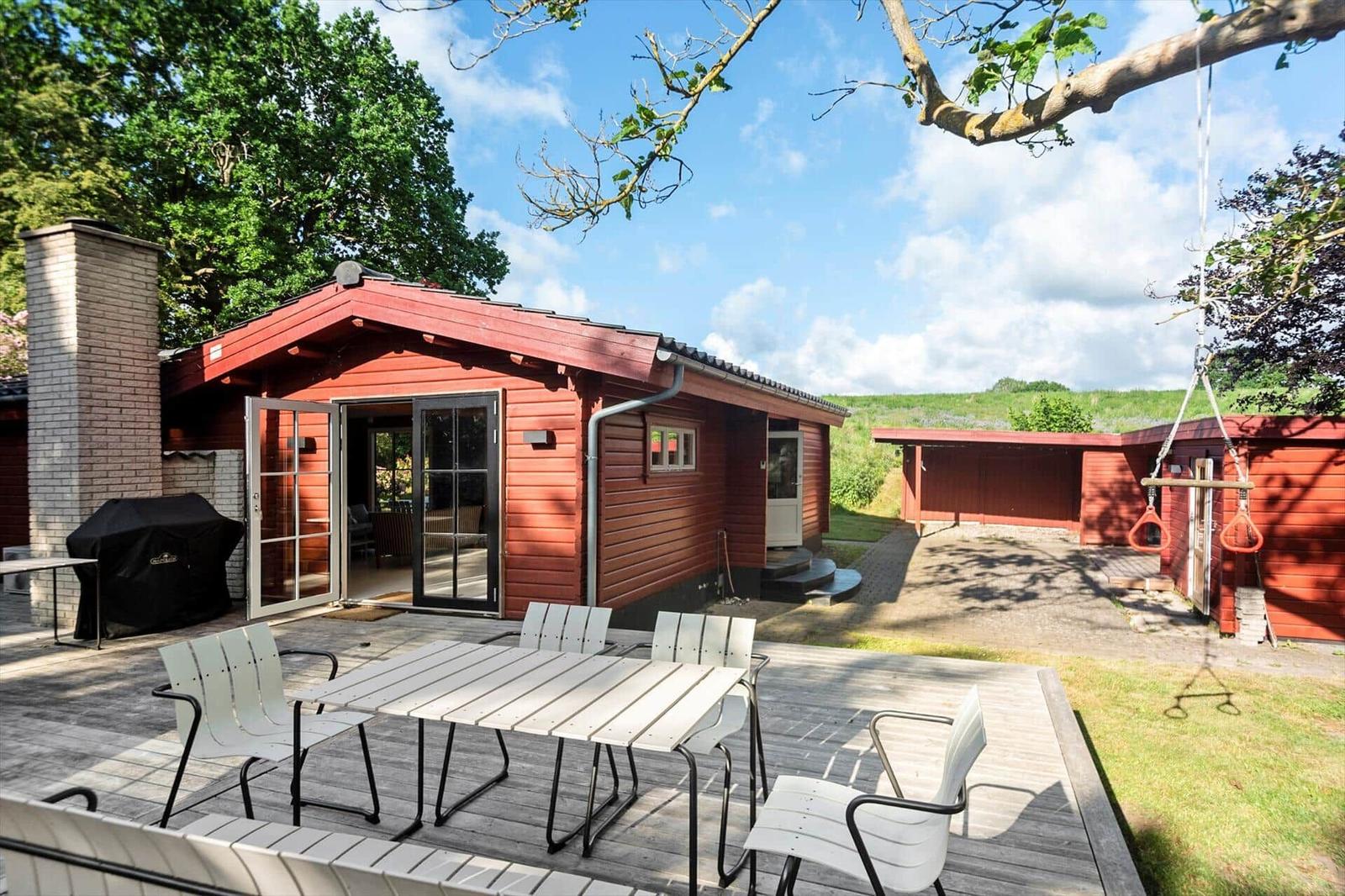 Roter Holzhaus mit Terrasse, Grill und Hängeseil. Grün hinter dem Haus.