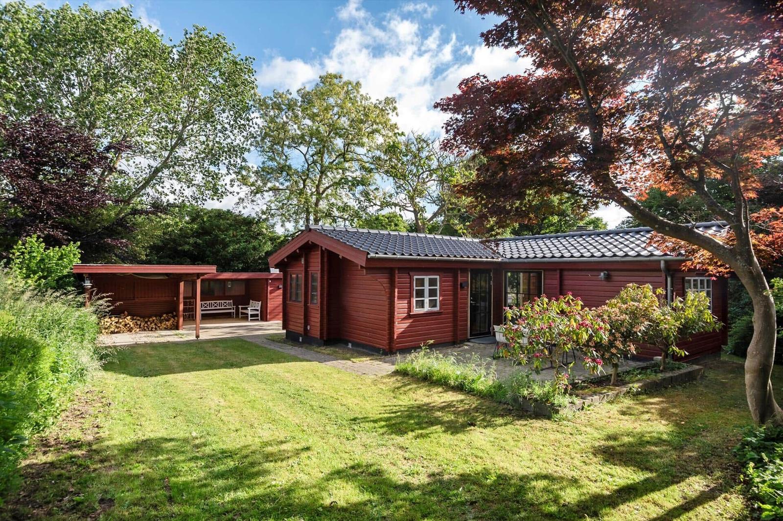 Rotes Holzhaus mit Garten, Terrasse und Grillplatz unter Bäumen.