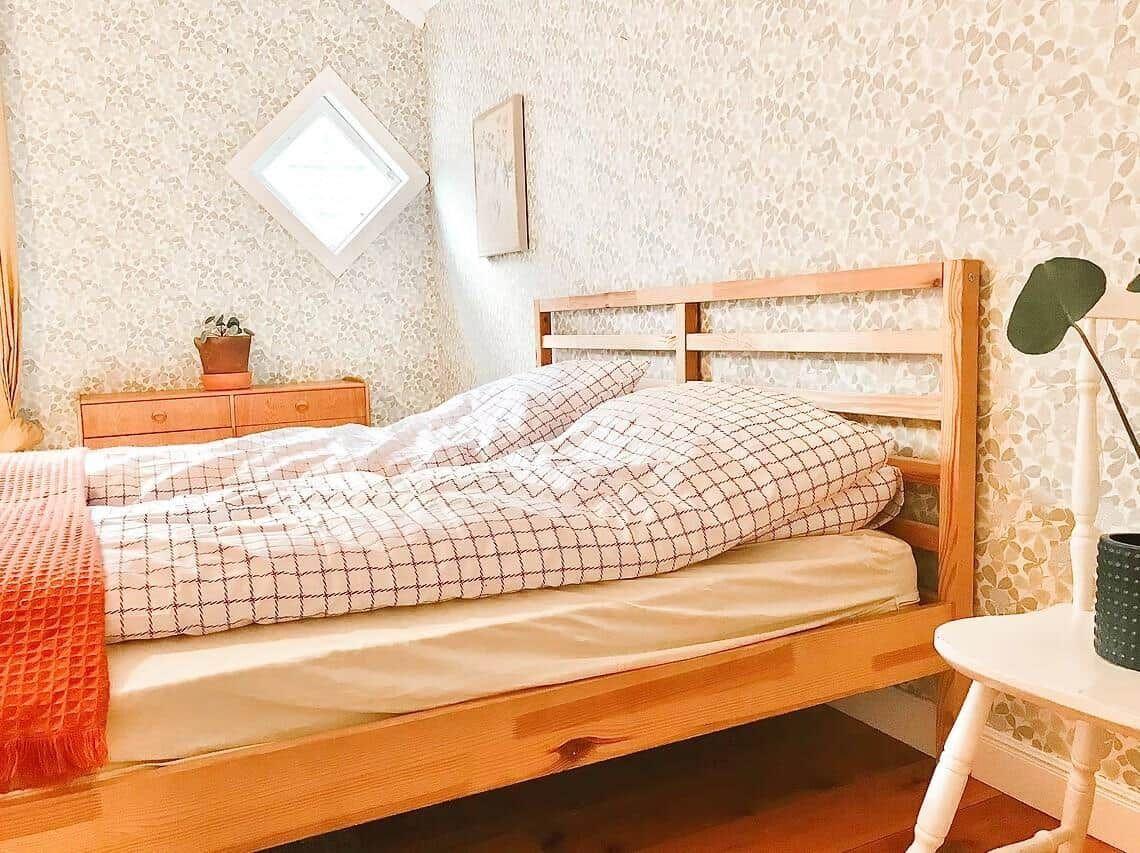 Ein Bett mit Holzgestell, gemustertem Bettbezug und einer roten Decke. Wand mit Tapete und kleinem Fenster.