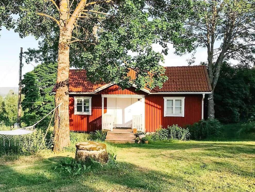 Rotes Holzhaus mit weißen Fenstern und Tür, umgeben von Bäumen und Gras.