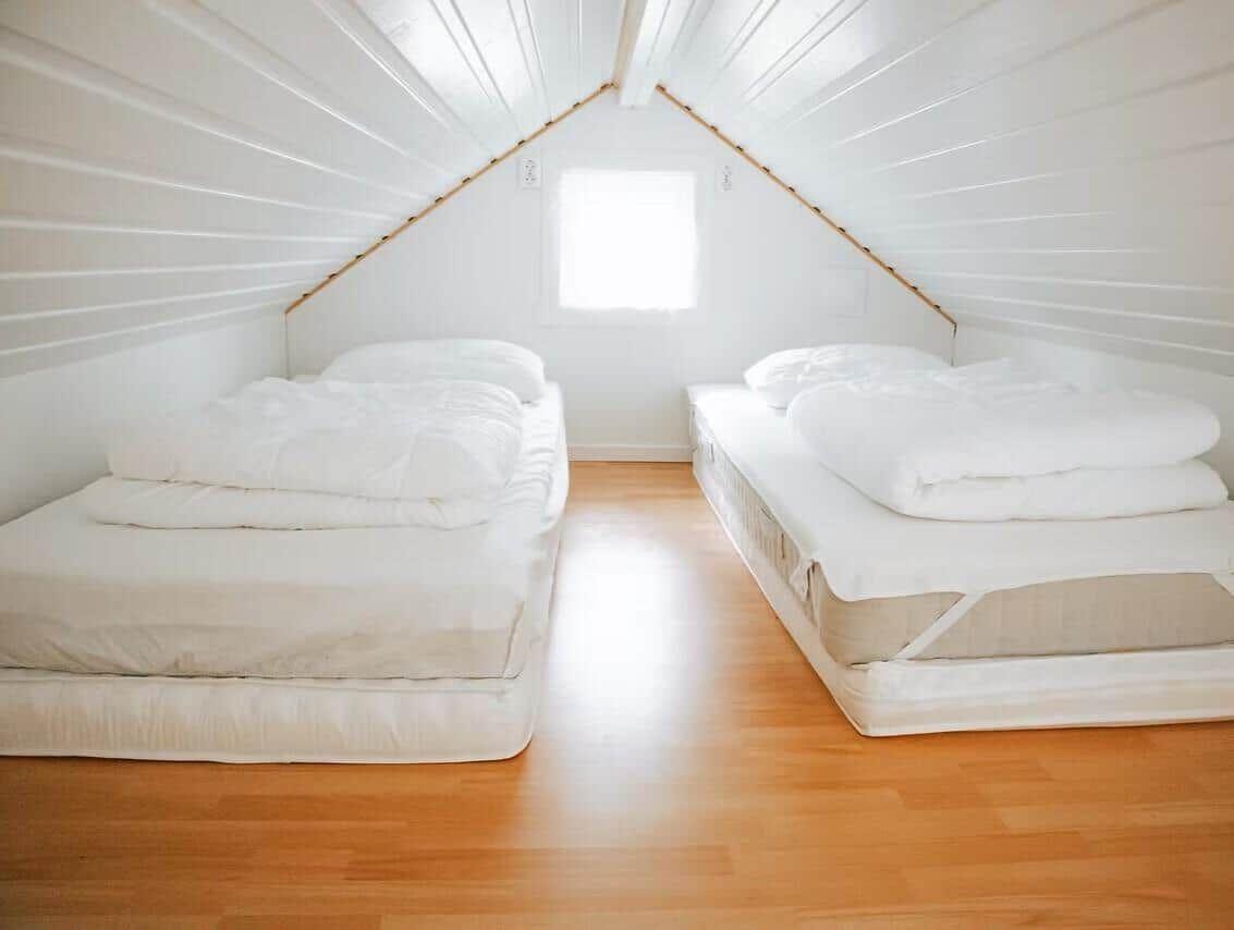 Zwei Matratzen mit weißen Bettwäschen in einem Dachbodenzimmer mit Holzboden und Fenster.