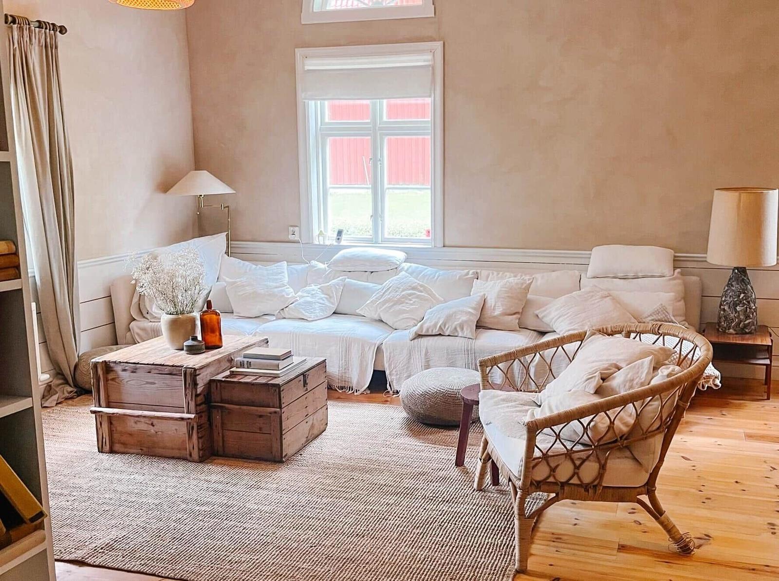 Ein Wohnzimmer mit Sofa, Rattanstuhl und Holztisch. Fenster mit Blick auf rotes Gebäude.