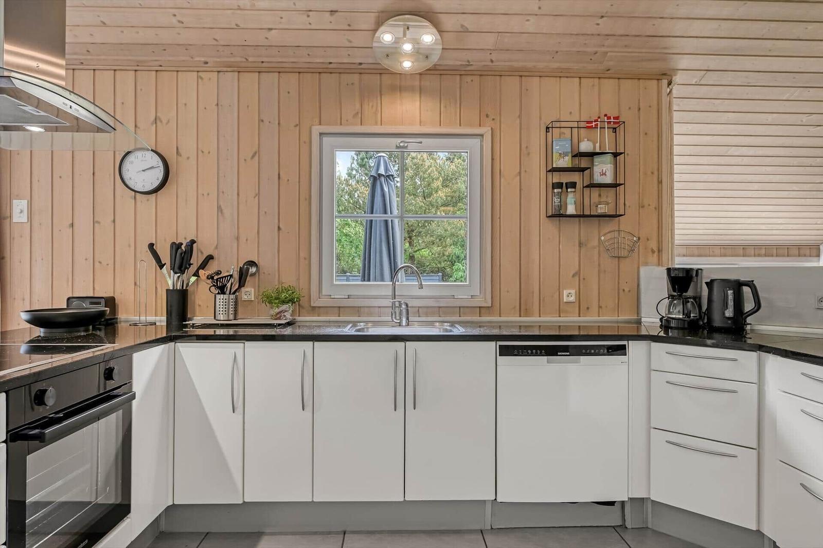Küche mit weißen Schränken, schwarzer Arbeitsplatte und Holzwand. Fenster mit Blick auf Garten.