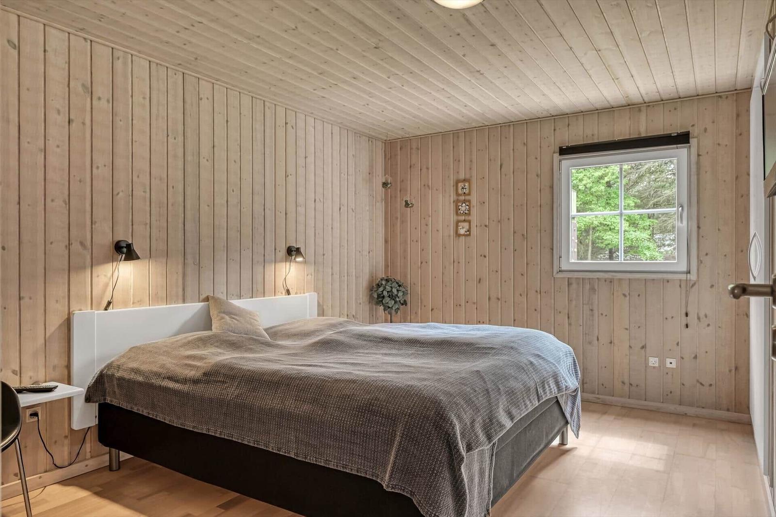 Schlafzimmer mit Holzwandverkleidung, Bett, Fenster und Nachttisch.