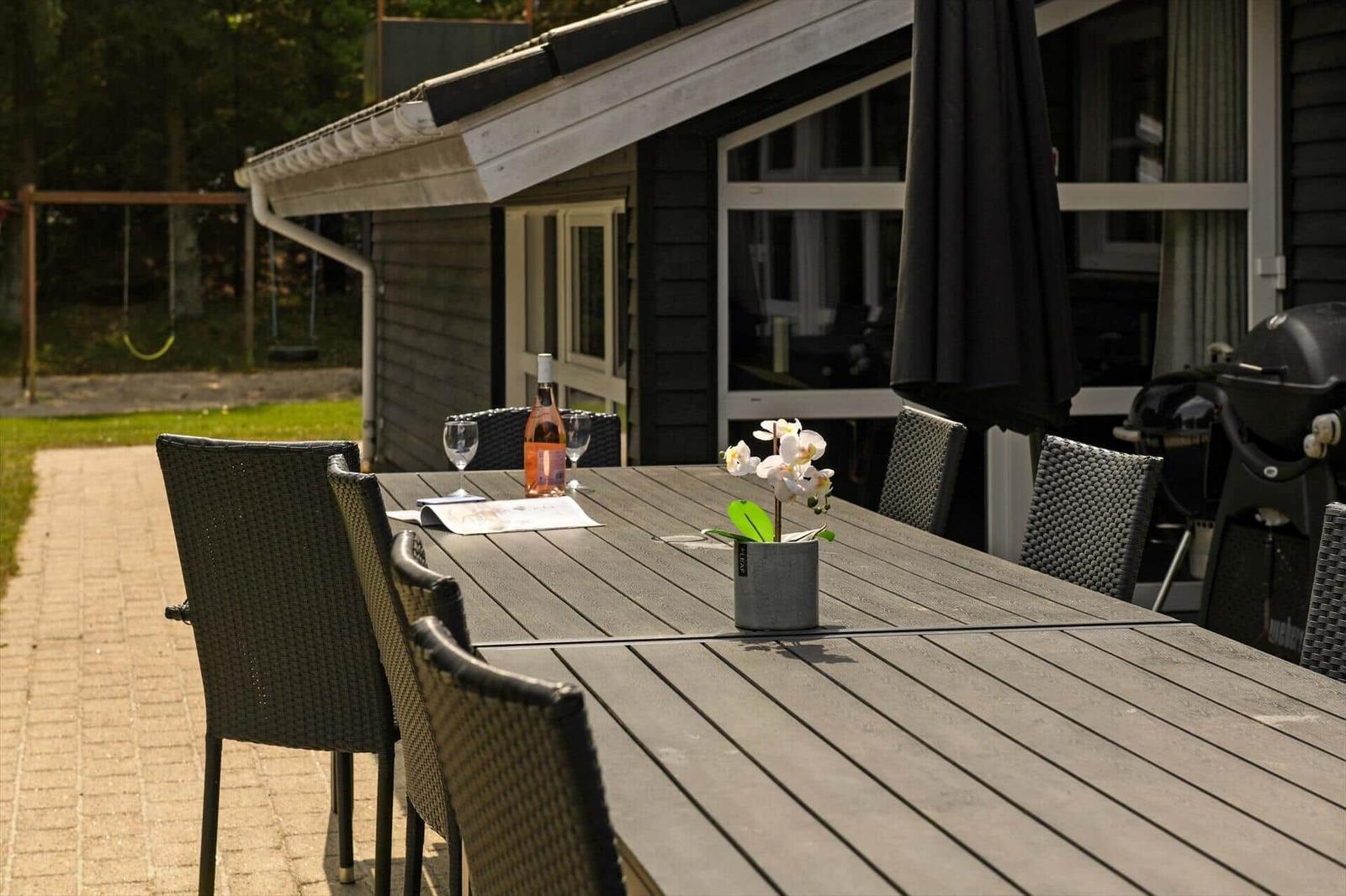 Außenbereich mit Tisch, Stühlen, Blumen und Grill. Hintergrund: Haus und Spielplatz.