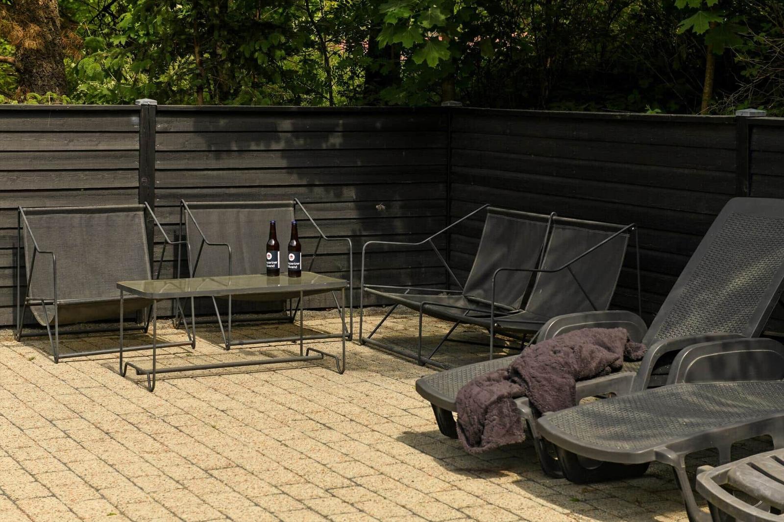 Terrasse mit Liegestühlen, Tisch und zwei Flaschen. Hintergrund: dunkler Zaun und Bäume.