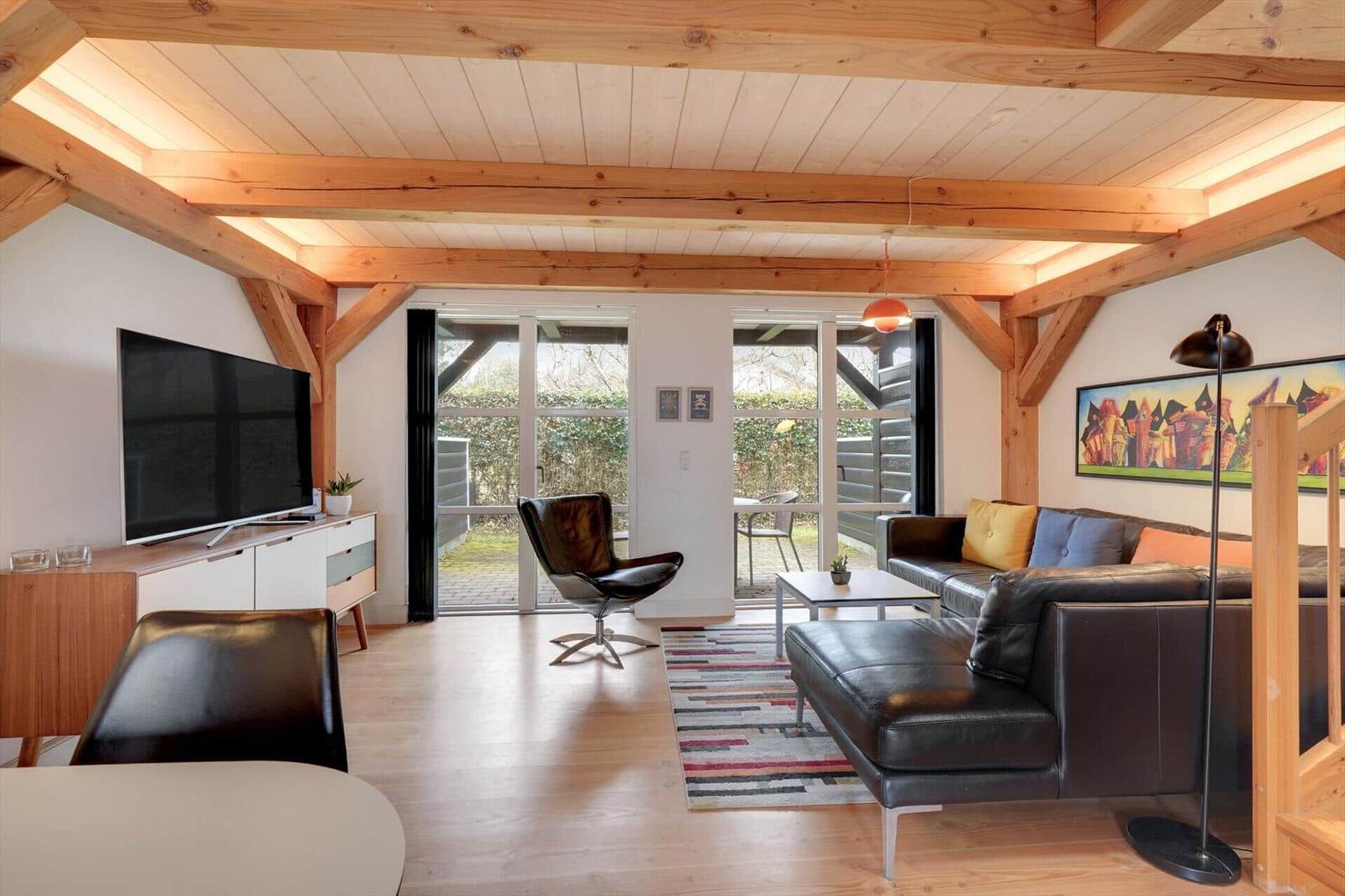 Wohnzimmer mit Holzbalkendecke, Sofa, Fernseher und Glastüren zum Garten.