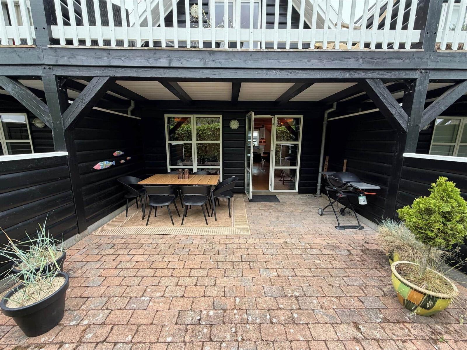 Terrasse mit Tisch, Stühlen, Grill und Pflanzen vor einem Haus mit Balkon.