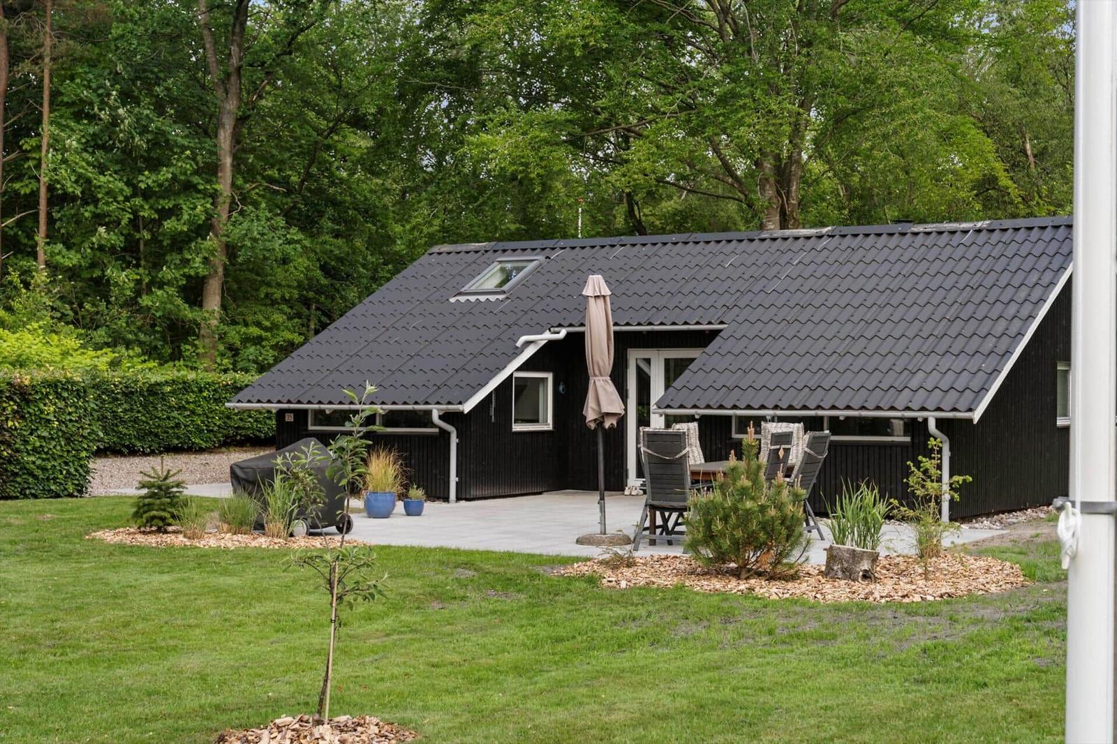 Schwarzes Ferienhaus mit Terrasse, Garten und Grillplatz. Umgeben von Bäumen.