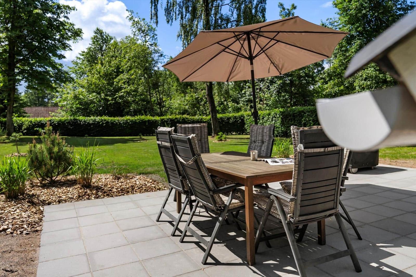 Terrasse mit Tisch, Stühlen und Sonnenschirm. Grünfläche und Bäume im Hintergrund.