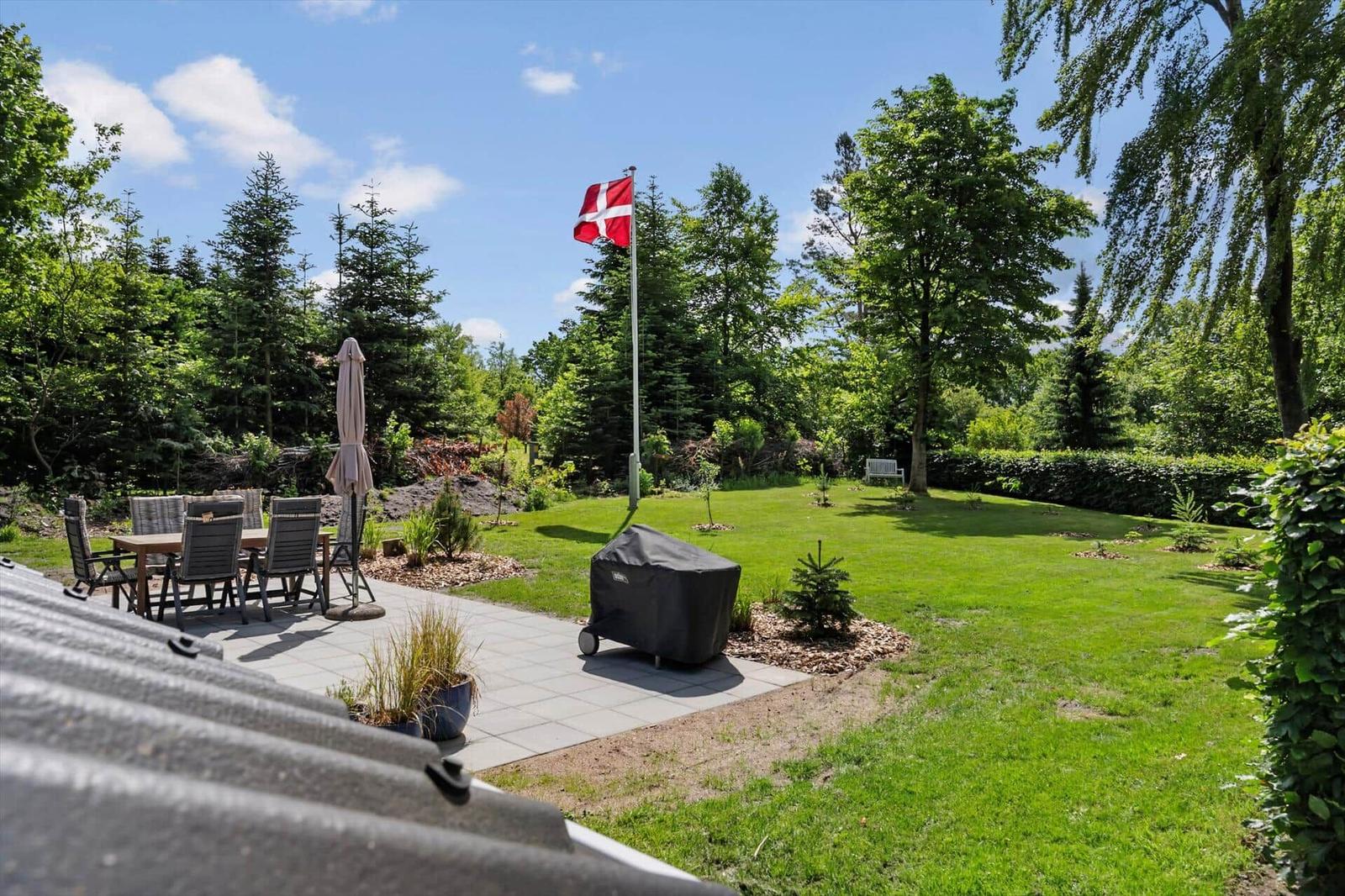 Garten mit Grill, Tisch, Stühlen und dänischer Flagge. Grünfläche und Bäume umgeben den Außenbereich.