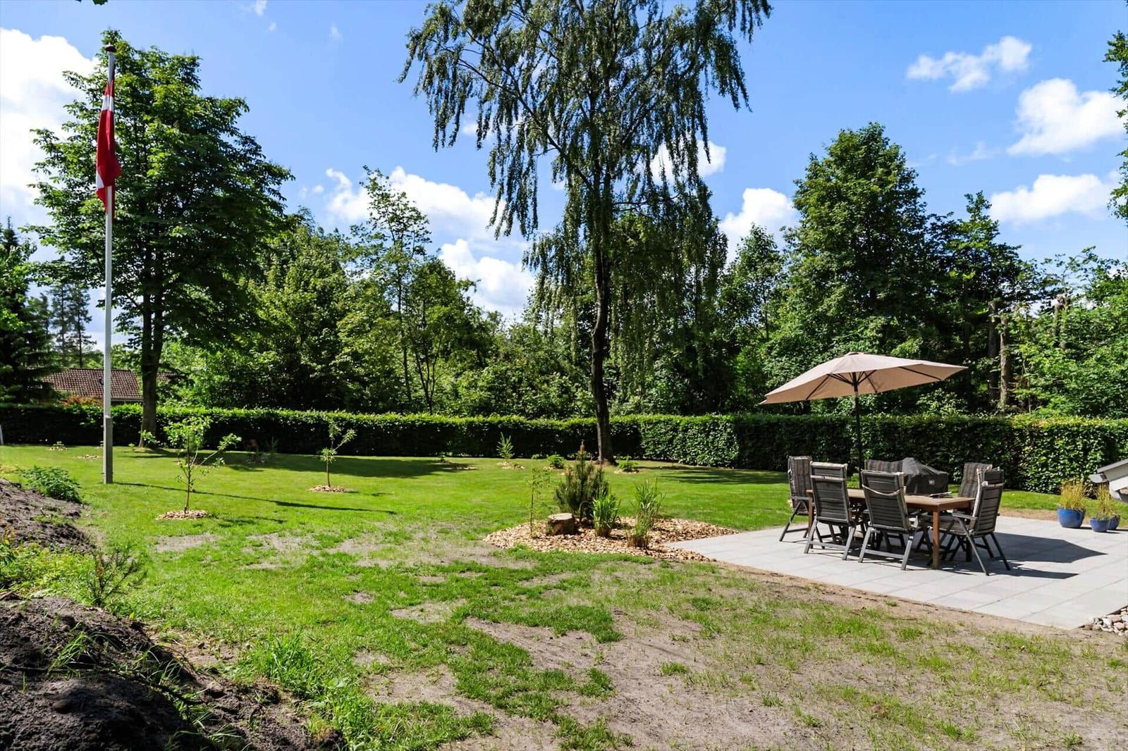 Hinterhof mit Grillplatz, Sitzgruppe unter Sonnenschutz und Flagge. Grünfläche und Bäume umgeben den Bereich.