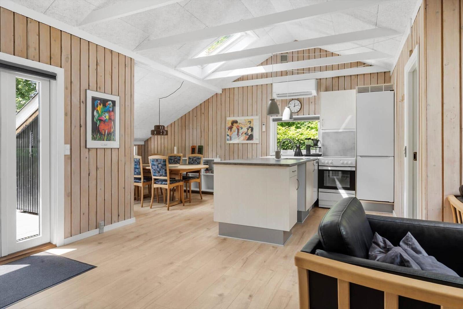Wohnküche mit Essbereich, Holzwänden und Dachschrägen. Schwarzleder-Sofa und Fenster mit Blick auf Grün.