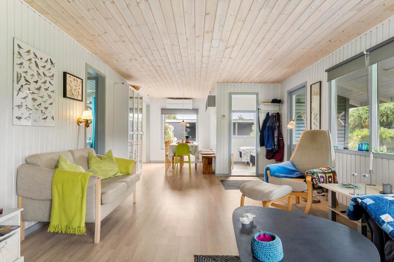 Wohnbereich mit Sofa, Sessel und Esstisch. Holzdecke und -boden. Fenster mit Blick auf Garten.