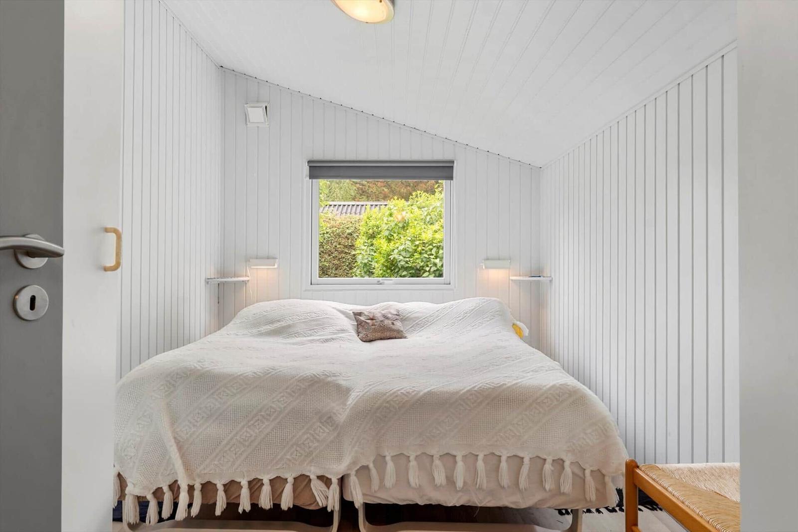 Ein Schlafzimmer mit weißem Bett, Wandpaneelen und Fenster zur Außenseite.