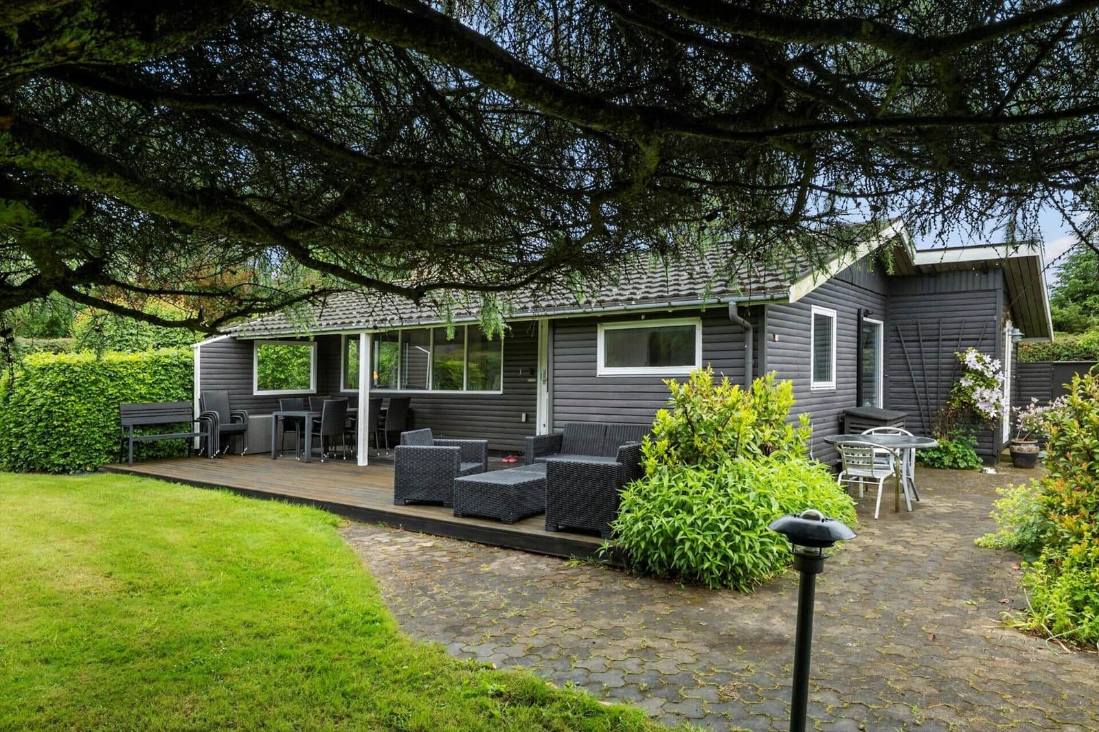 Gartenhaus mit Terrasse, Sitzgelegenheiten und Bäumen. Grünfläche und Pflanzen umgeben das Haus.