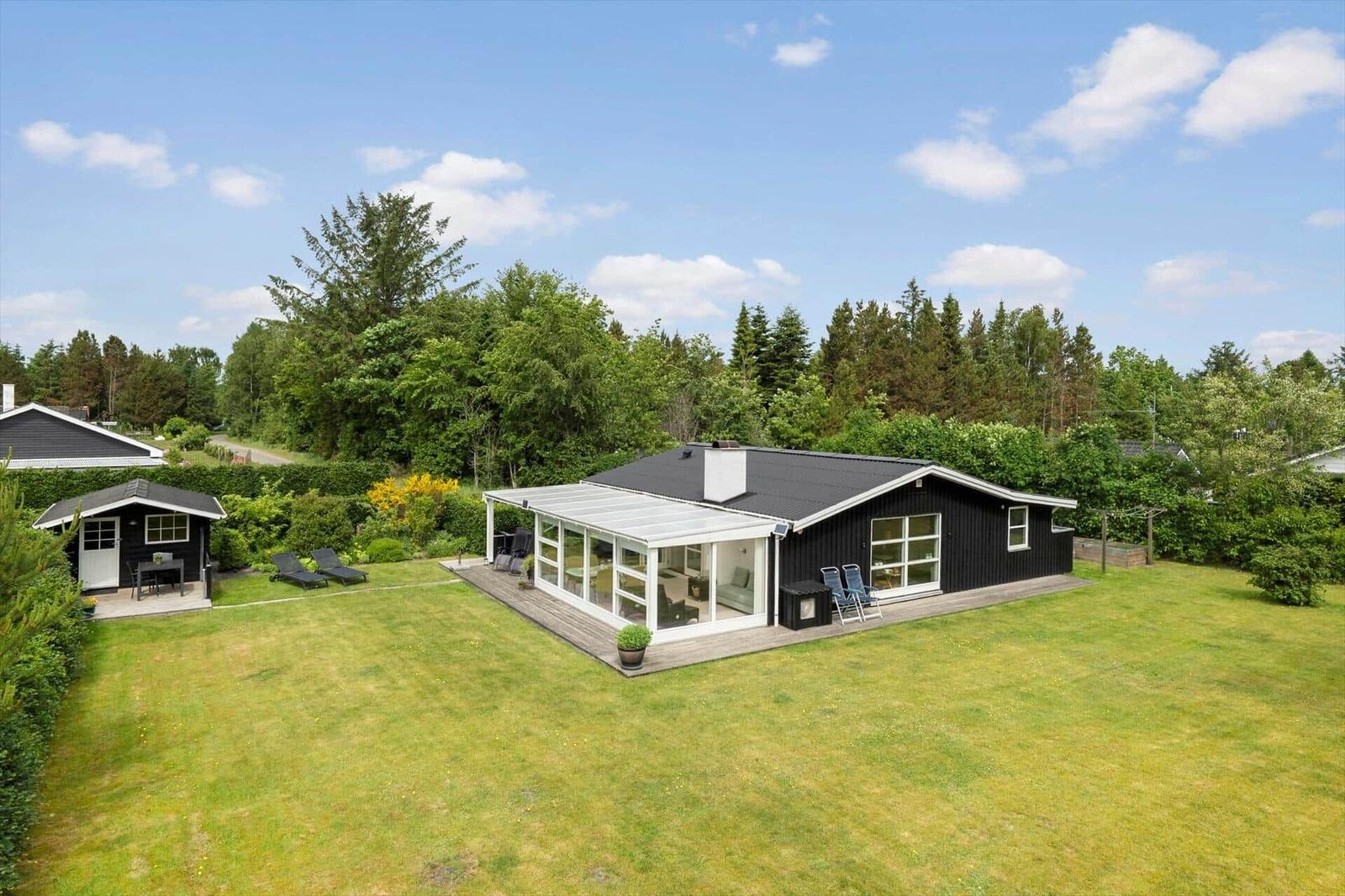Schwarzes Haus mit Glasveranda, Garten und Liegestühlen. Umgeben von Bäumen und Grün.