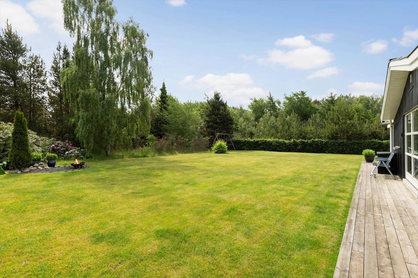 Großer Garten mit Rasenfläche, Holzterrasse und Feuerkorb. Umgeben von Bäumen und Sträuchern.