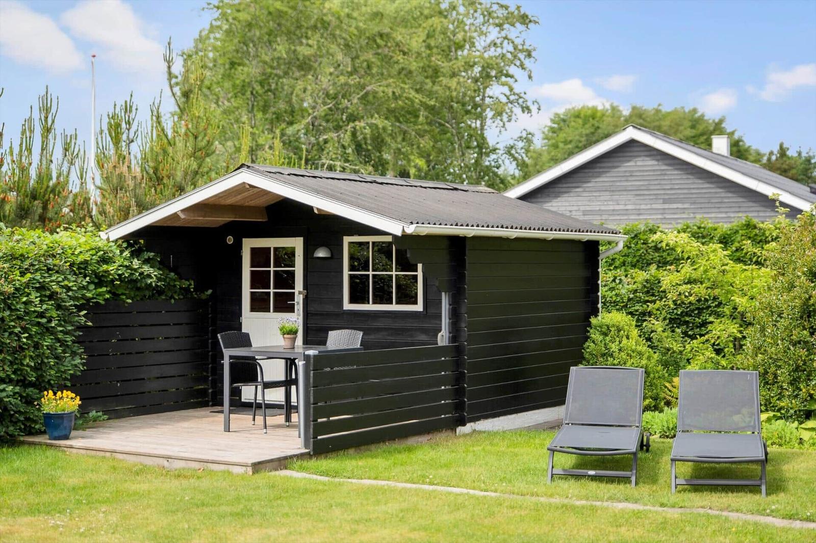 Schwarze Hütte mit Terrasse, Tisch, Stühlen und Liegestühlen im Garten.