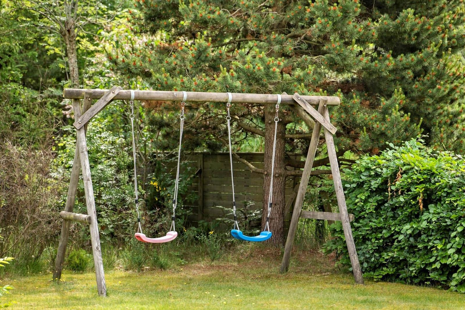 Holzschaukel mit zwei Sitzplätzen im Garten. Hintergrund: Bäume und Sträucher.