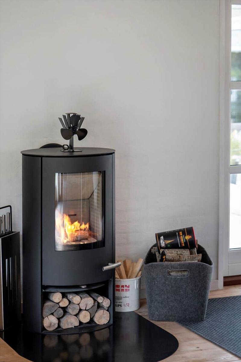 Schwarzer Holzofen mit brennendem Feuer, darunter Holzstapel und daneben Korb mit Brennholz.