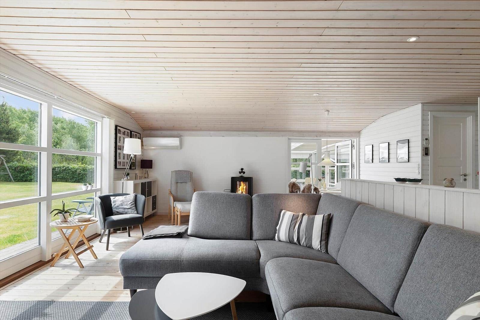 Wohnzimmer mit grauem Sofa, Kamin und großen Fenstern zur grünen Außenansicht.