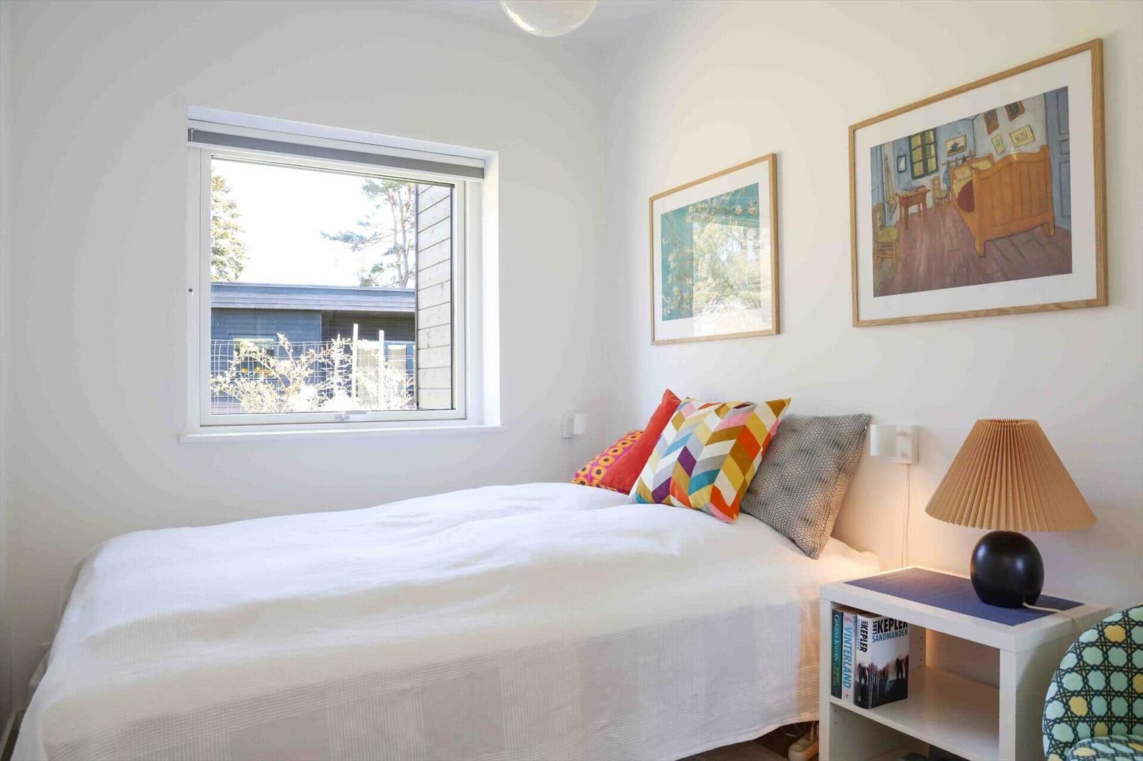 Bedroom with bed, window, and wall art. Nightstand with lamp and books.