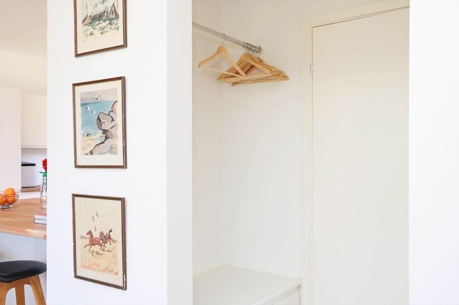 Closet with wooden rod and hooks. Wall with three framed pictures. View into kitchen area.
