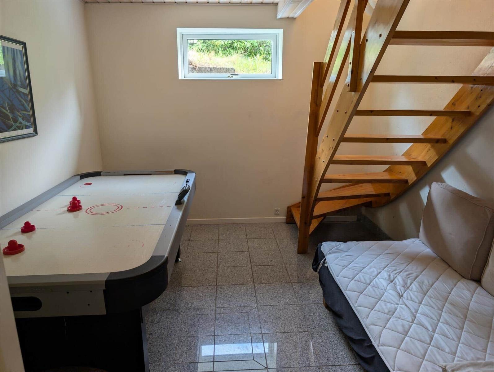 Room with air hockey table, sofa bed, and wooden staircase. Window with view of green area.