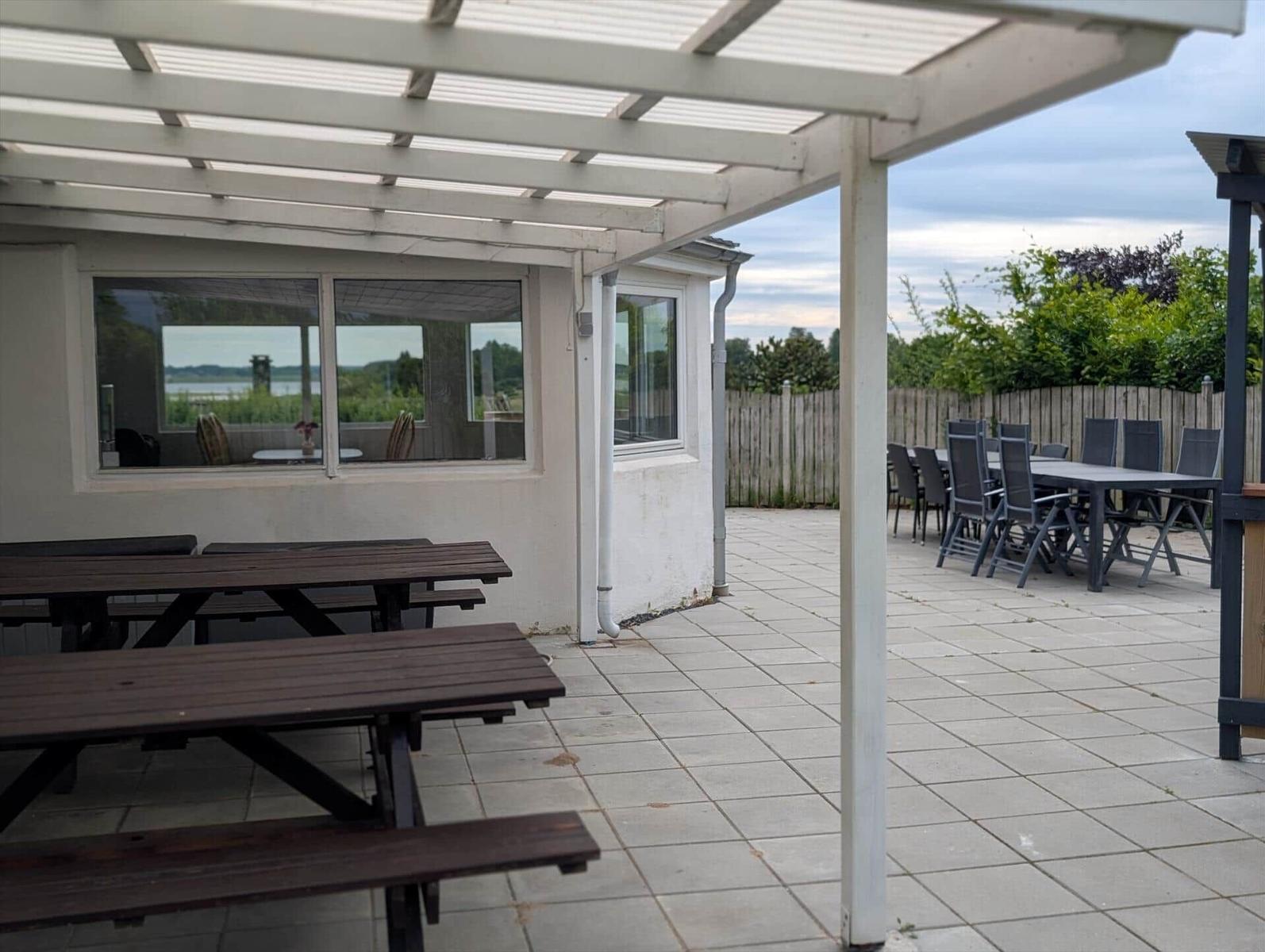 Terrace with tables and chairs under a roof. View of a lake through windows.