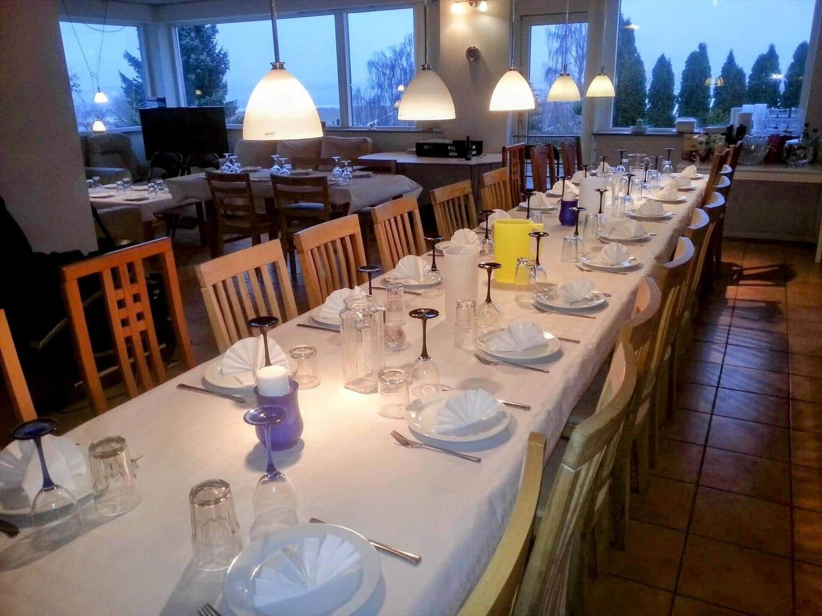 Long table with chairs, tablecloth, dishes, and glasses. Windows with view of trees.