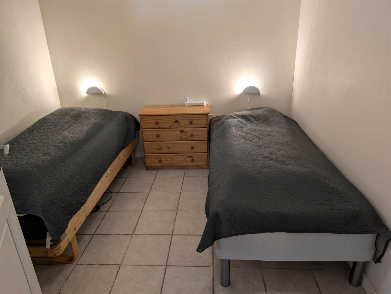 Two single beds with gray bedding and wooden dresser in a room with tiled floor.