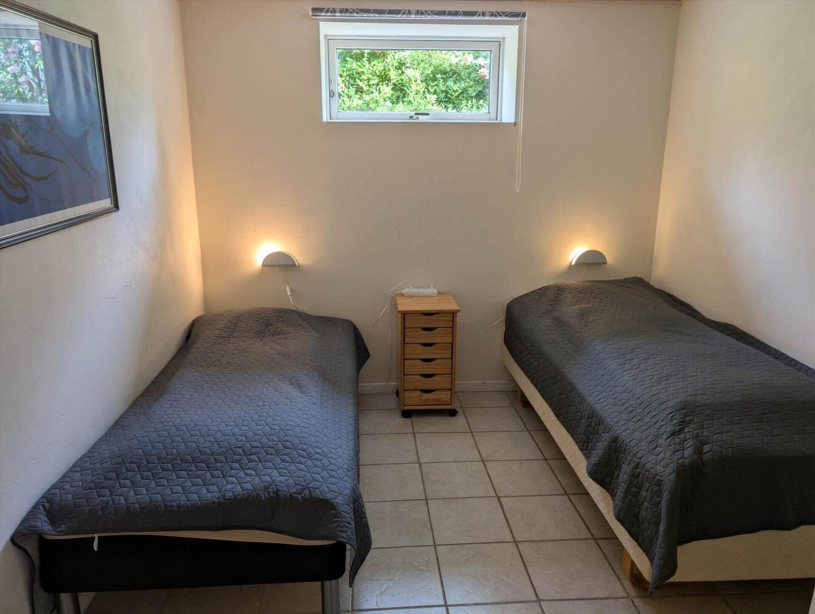 Two beds with gray covers, a small cabinet, and a window with a view of greenery.