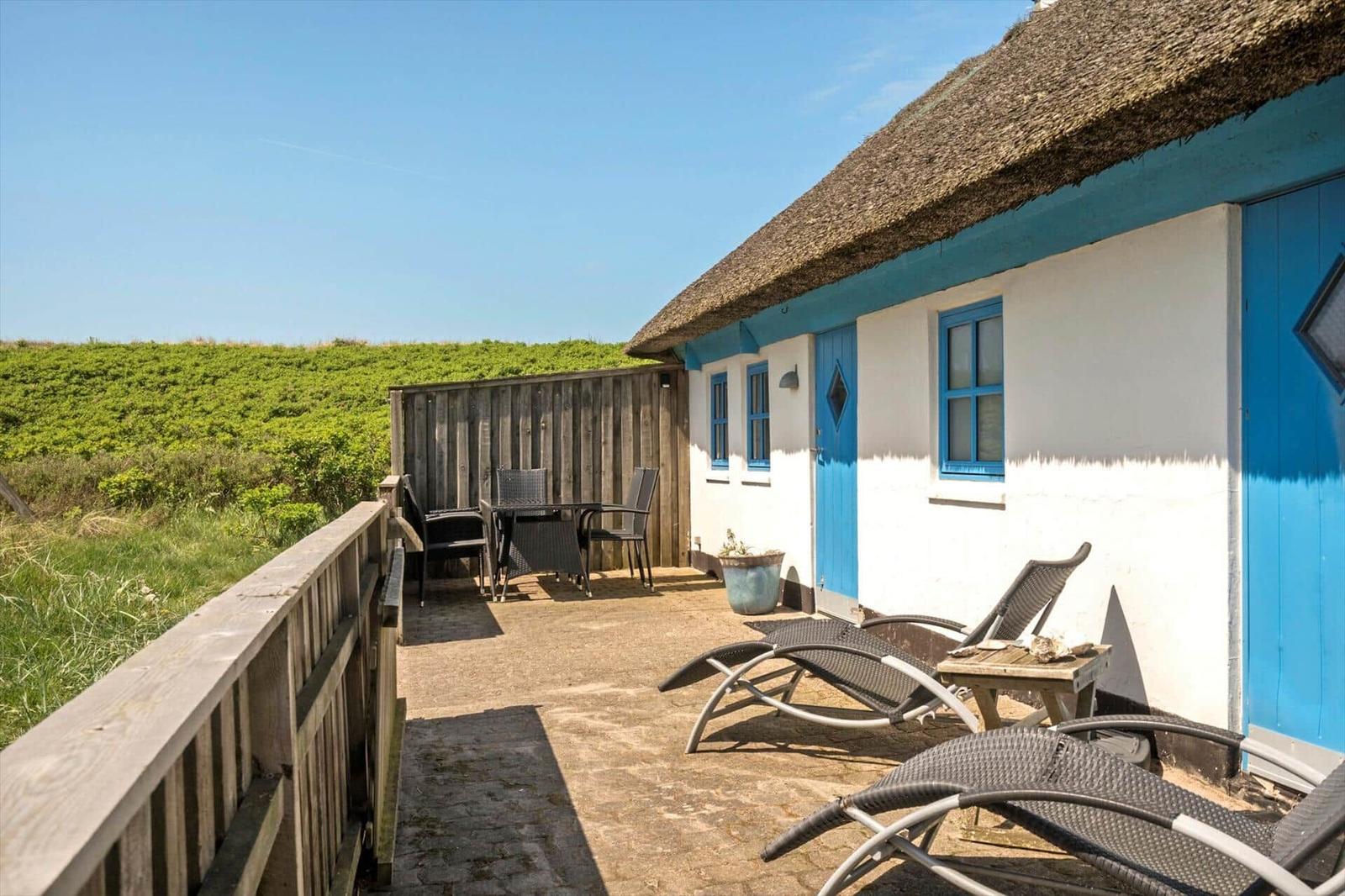 Terrasse mit Liegestühlen und Tisch vor einem blauen Haus mit Strohdach.