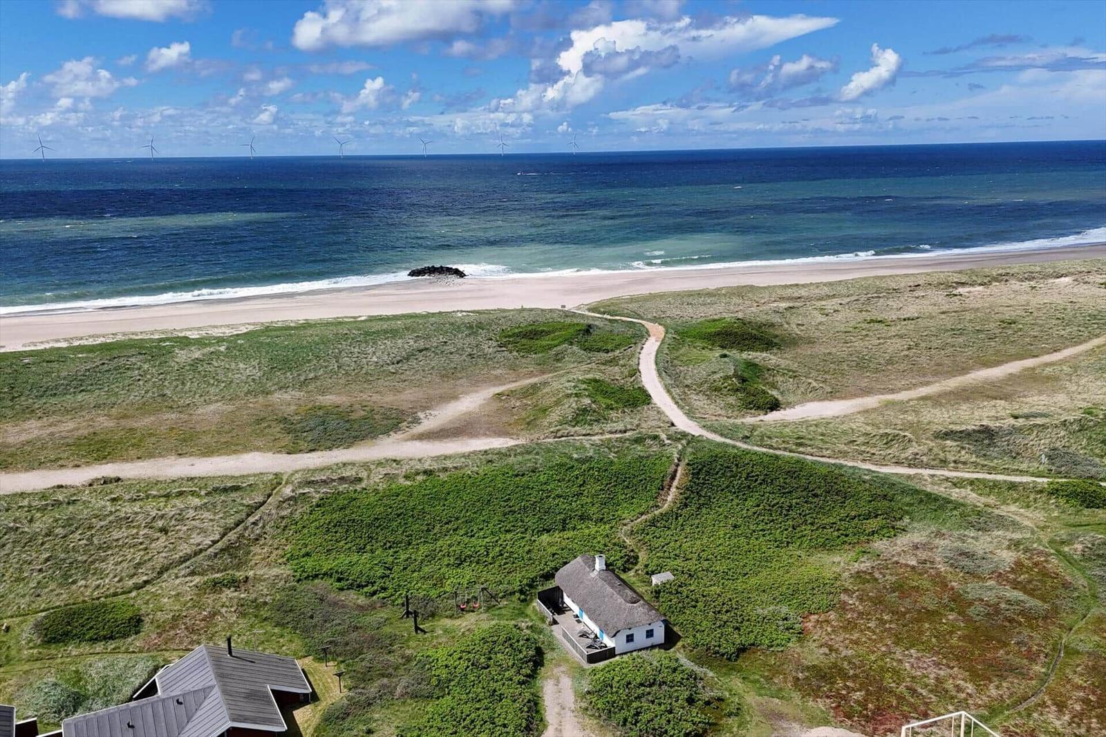 Haus mit Dach aus Stroh, umgeben von Grasland und Strand. Im Hintergrund sind Windräder und das Meer.