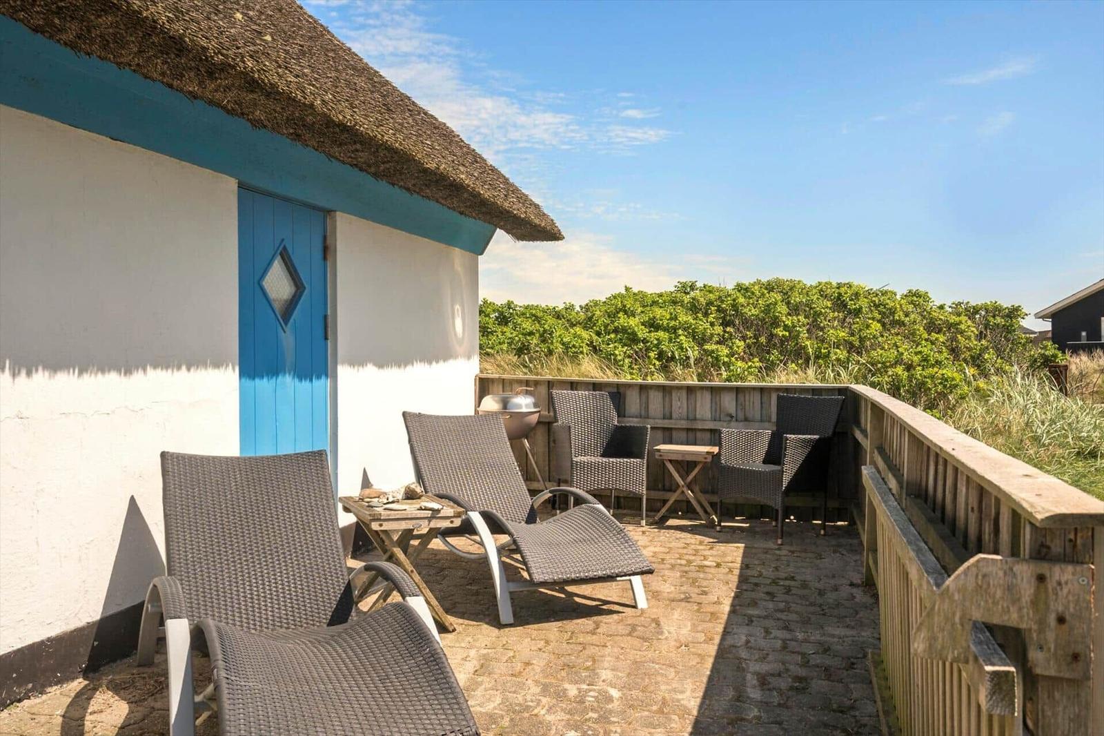 Terrasse mit Liegestühlen, Tisch und Grill vor einem Haus mit Strohdach und blauer Tür.