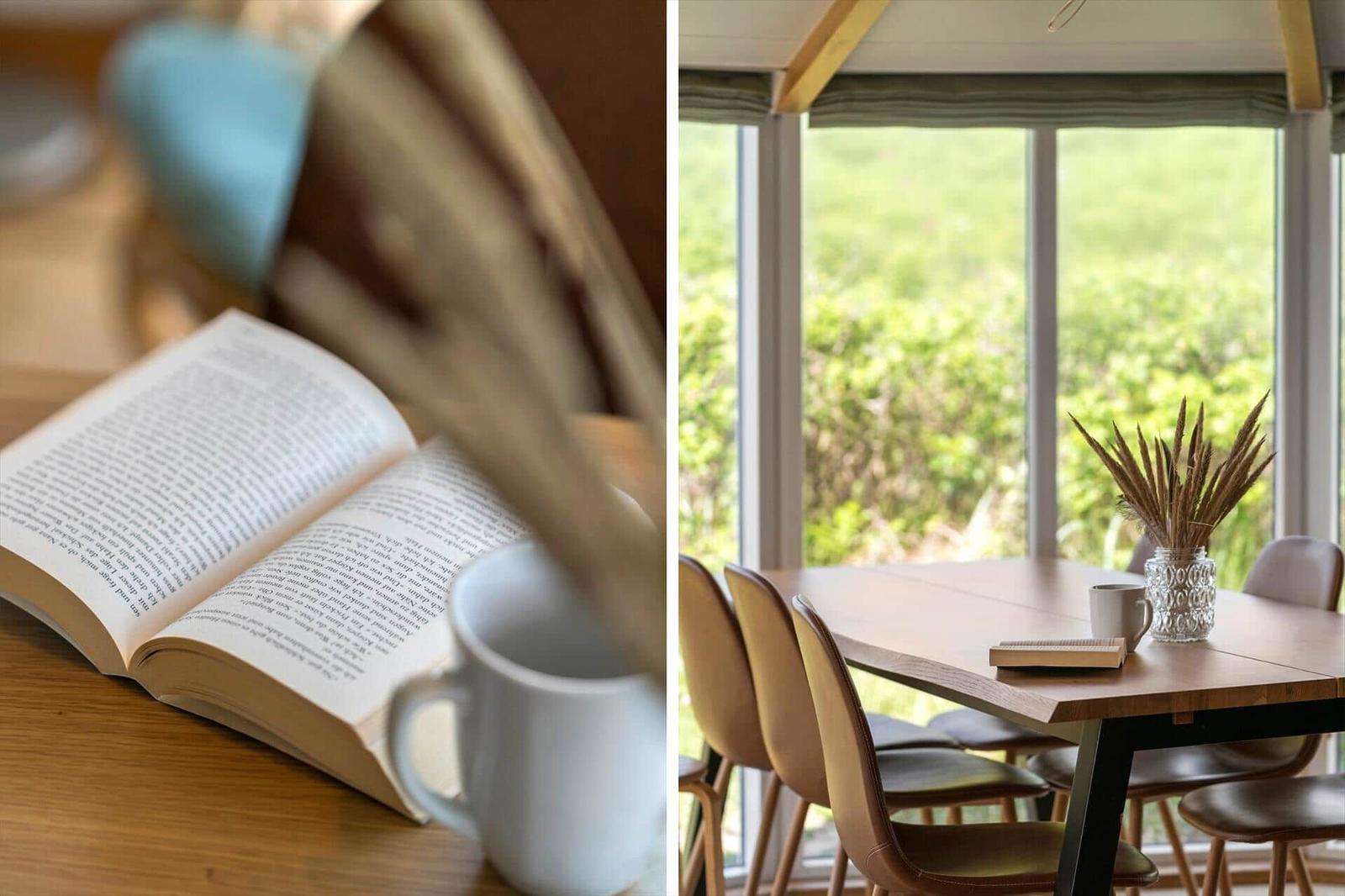 Ein Tisch mit Stühlen und einem Fensterblick auf Grün. Ein Buch und eine Tasse auf einem Holztisch.