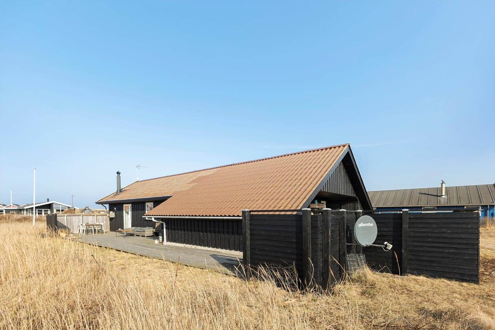 Ein dunkles Holzhaus mit braunem Dach und Terrasse. Umgeben von trockenem Gras und einem Zaun.