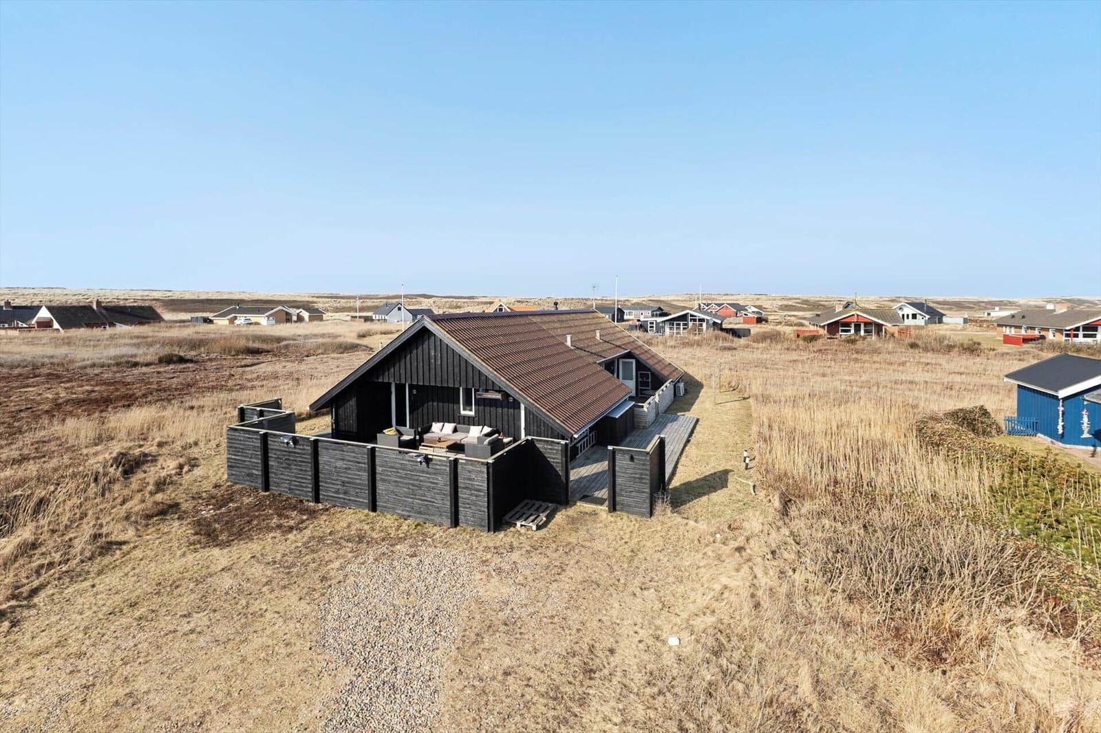 Schwarzes Holzhaus mit Terrasse und Garten, umgeben von trockenem Gras und weiteren Häusern.