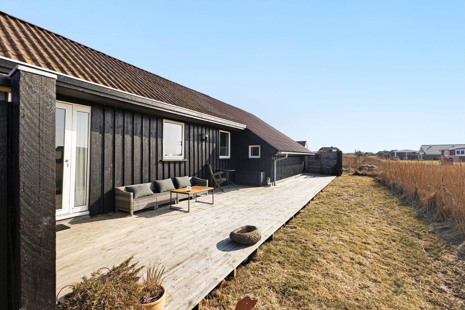 Holzterrasse mit Sitzmöbeln vor einem dunklen Holzhaus mit Schiebetür und Fenstern.