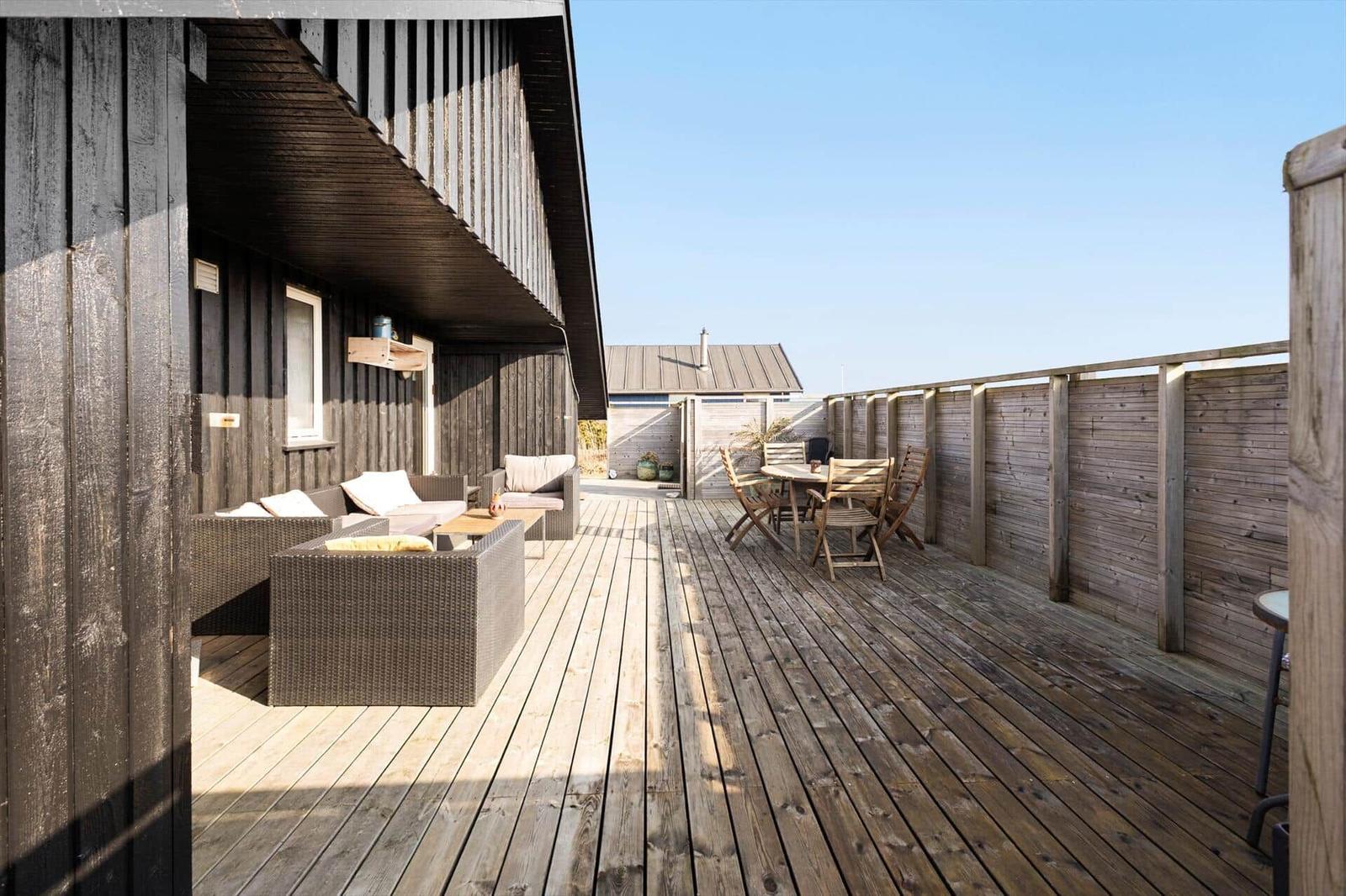 Holzterrasse mit Sitzgelegenheiten und Tisch. Holzwand und Himmel sind sichtbar.