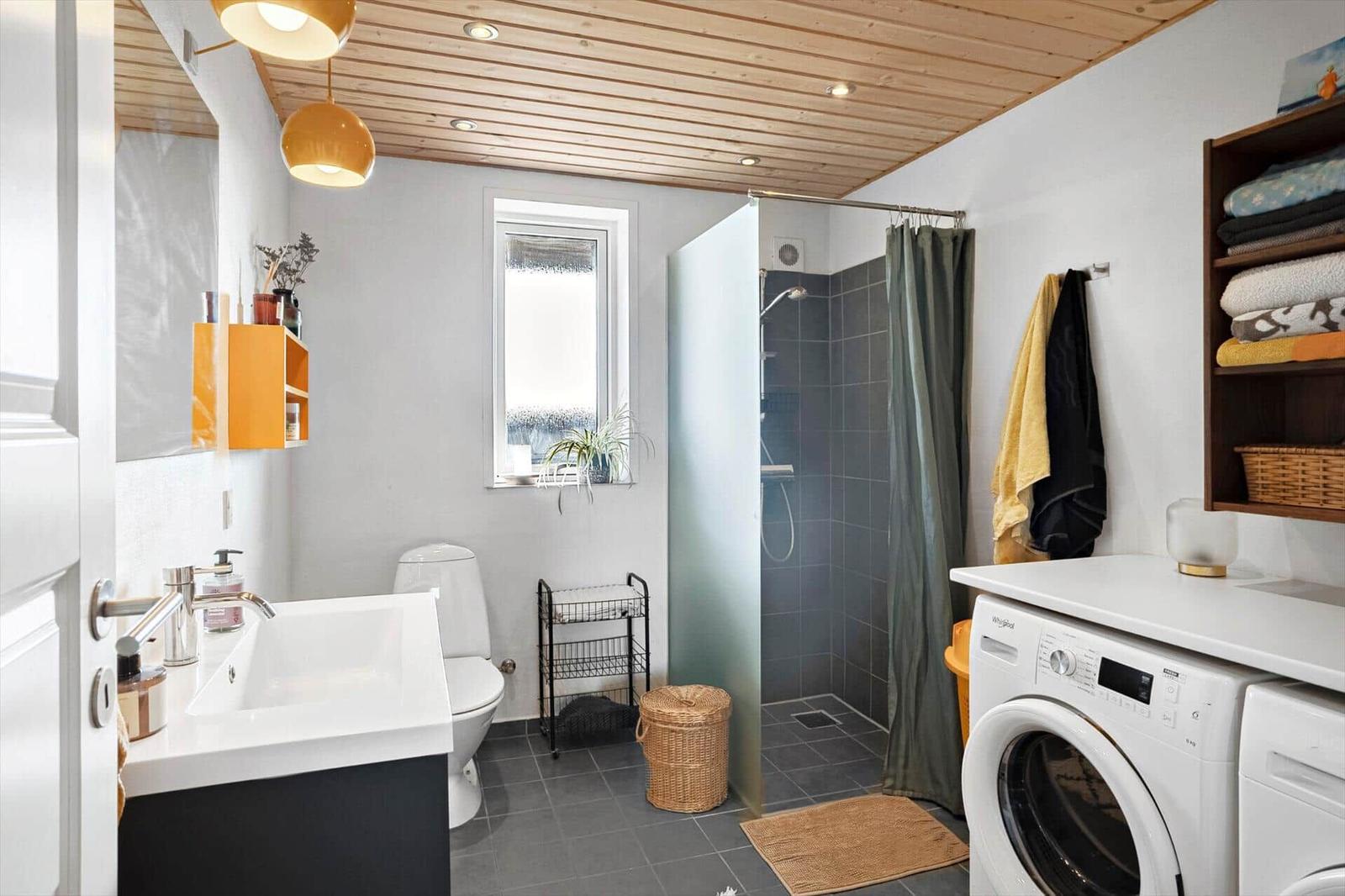 Badezimmer mit Dusche, Waschmaschine und Waschbecken. Holzdecke und Fenster.