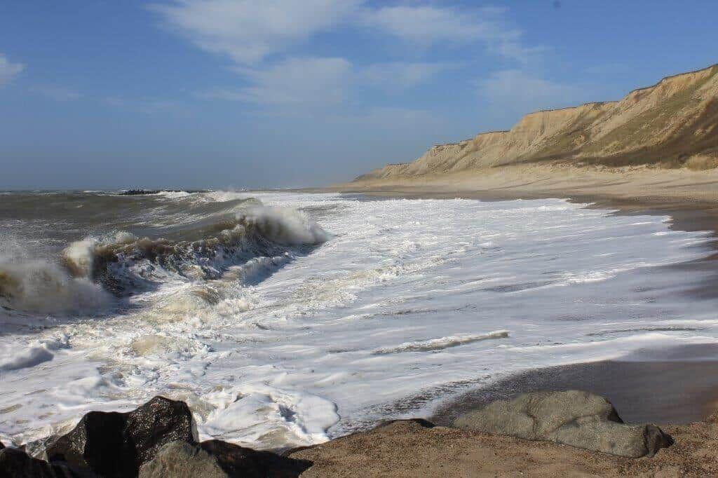 Wellen brechen an Strand mit Felsformationen im Vordergrund und steilen Uferhängen im Hintergrund.