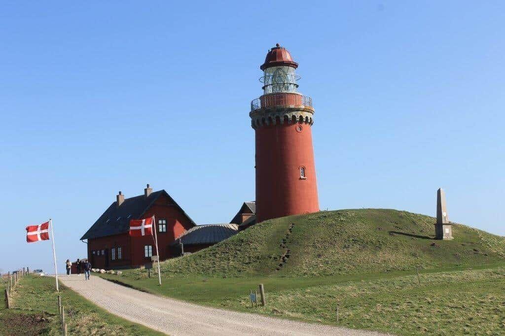 Roter Leuchtturm auf grüner Anhöhe mit daneben stehendem Haus und dänischen Flaggen.