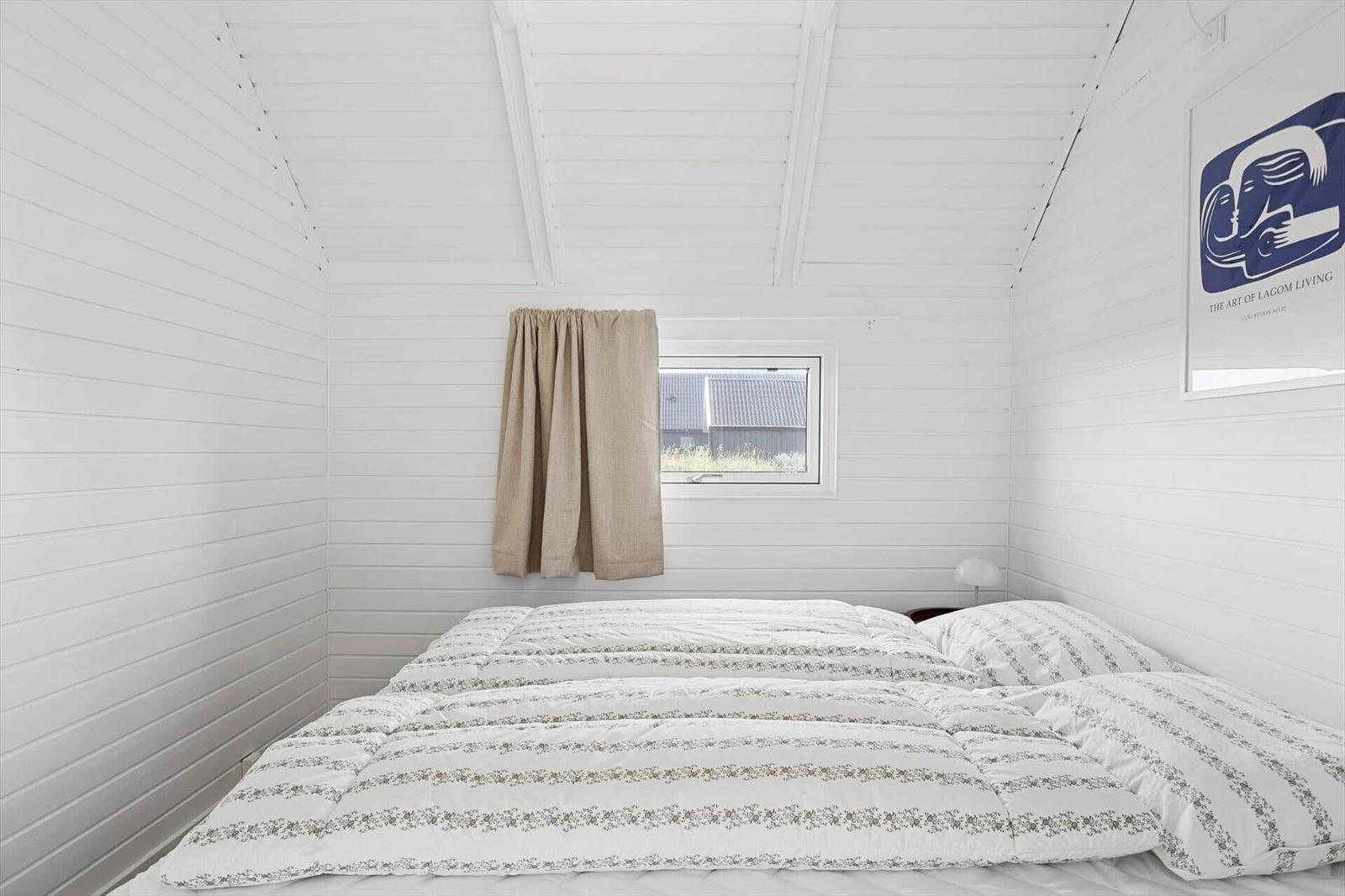 Bedroom with white wood paneling, bed, and small window with curtain.