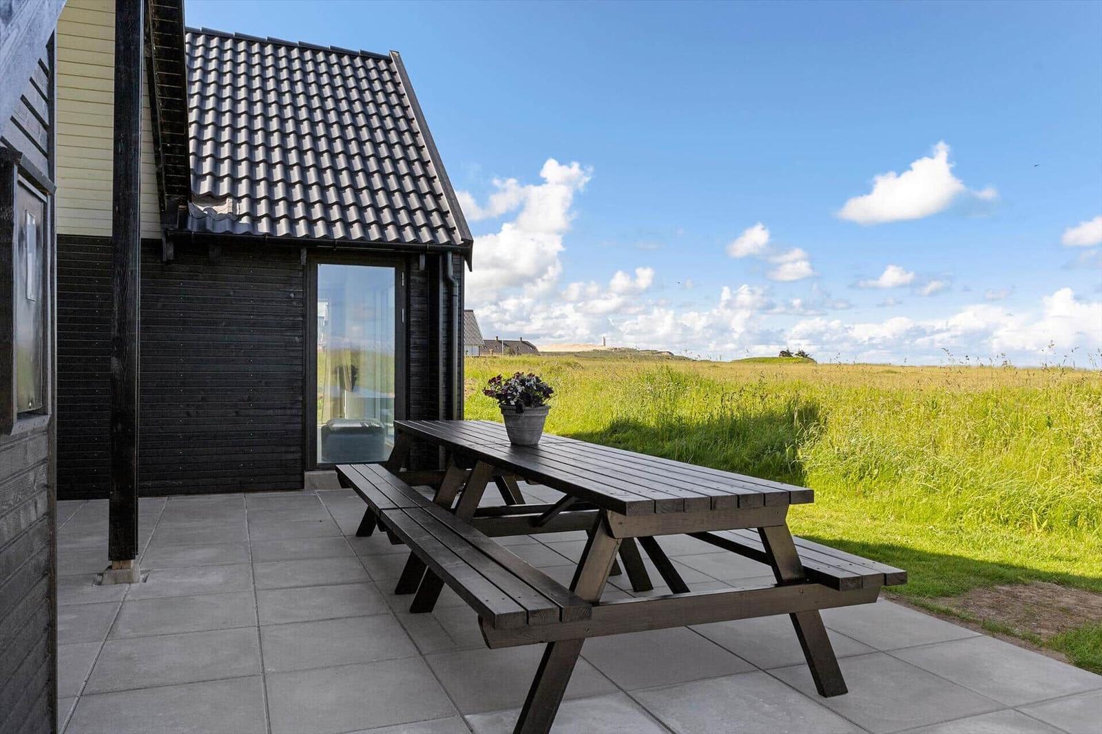 Terrace with wooden table and benches in front of a house with roof and glass facade.