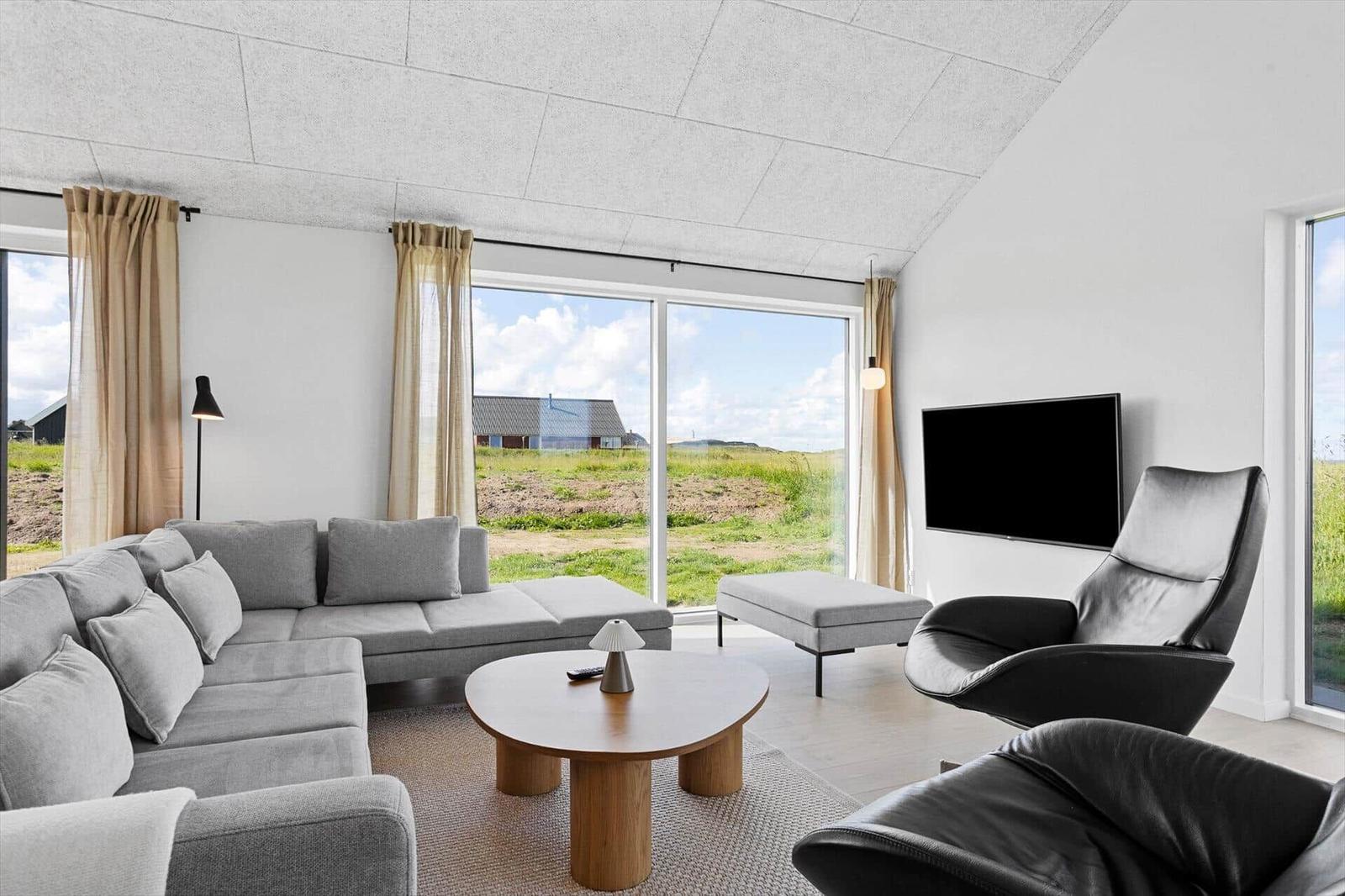 Living room with sofa, chairs, and TV. Large windows show landscape.
