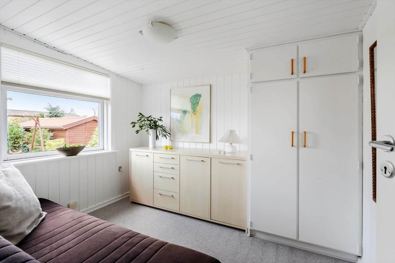 Schlafzimmer mit Fenster, Schrank und Schreibtisch. Draußen ist ein Garten mit Haus.